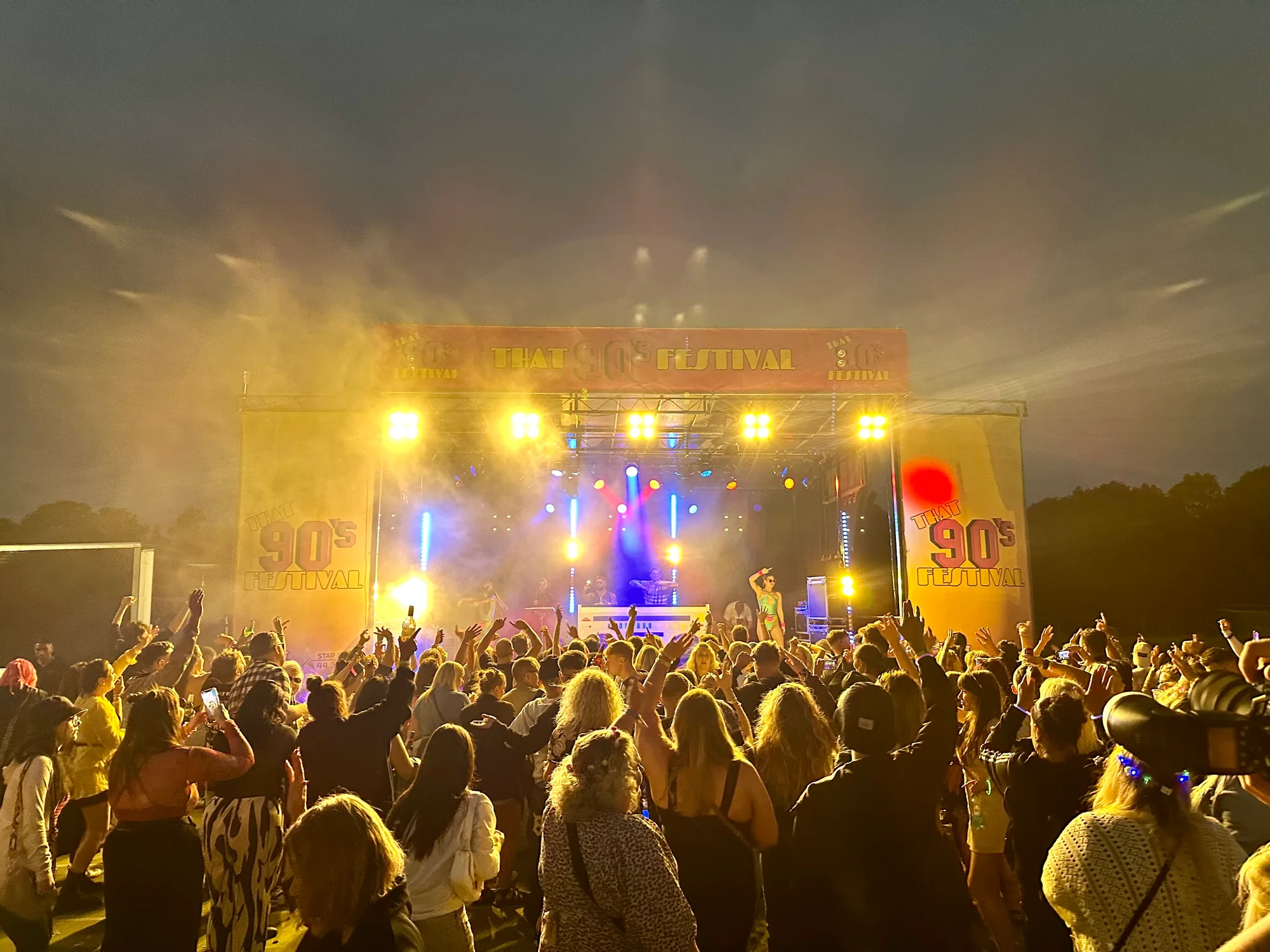Crowd watching the fully branded That's 90s Festival stage with bright show lighting in Corby
